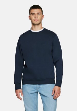 Homme aux cheveux courts et bruns portant un sweat-shirt bleu marine à col rond et un jean bleu clair, debout devant un fond uni.