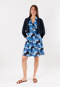 Abito blu e nero con motivo tie-dye, caratterizzato da una vita stretta e un elemento decorativo circolare; abbinato a una giacca navy corta e sandali marroni.