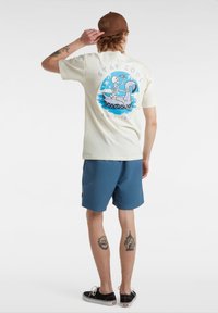 Helles Cremet-Shirt mit einem bunten Grafikdesign, das ein Skelett im Kajak zeigt. Kombiniert mit blauen Shorts und schwarzen Schuhen. Braune Mütze mit Logo.
