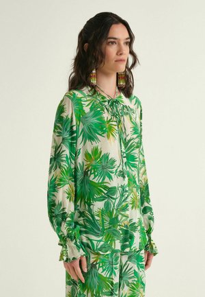 Woman with dark hair wears a green and white tropical leaf print blouse and matching pants, accessorized with long multicolored beaded earrings.