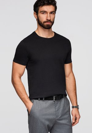 Mężczyzna z brodą, ubrany w czarny t-shirt i szare spodnie, stojący z jedną ręką w kieszeni na jednolitym tle.