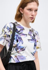 Γυναίκα που φοράει λευκό μπλουζάκι με μεγάλο μωβ λουλουδάτο σχέδιο και παγιέτες, μαύρο παντελόνι, ασημένια κρεμαστά σκουλαρίκια και μαύρη δερμάτινη τσάντα ώμου.