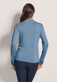 Frau in einem hellblauen Langarmoberteil mit pailettenbesetzten Bündchen und navyfarbener Hose, die mit dem Rücken zu einer schlichten Hintergrundkulisse steht.