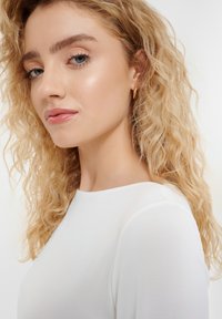 Zbliżenie osoby z falującymi, blond włosami, noszącej białą bluzkę. Wyróżniające się szczegóły to gładka tekstura i okrągły dekolt. Złote kolczyki w kształcie kółek.