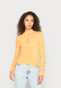 Top ajustado de mangas longas em tecido amarelo mostarda macio e canelado. Apresenta um recorte em formato de buraco de chave e detalhes drapeados na parte central da frente.