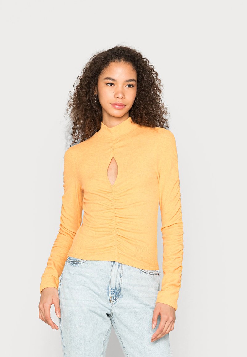 Top ajustado de mangas longas em tecido amarelo mostarda macio e canelado. Apresenta um recorte em formato de buraco de chave e detalhes drapeados na parte central da frente.
