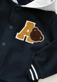 Dunkelblaues Sweatshirt mit grauer Kapuze, ausgestattet mit einem großen goldenen "A"-Aufnäher und einem braunen Bärenpatch, schwarzen Knöpfen und gestreiften Rippbündchen.