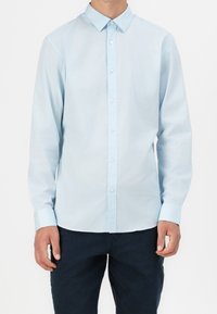 Homme portant une chemise bleu clair à manches longues boutonnée et un pantalon foncé, debout devant un fond blanc.