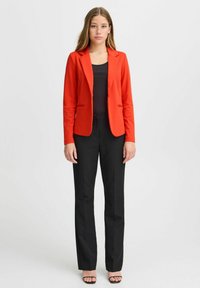 Blazer rouge au design ajusté avec deux poches avant, associé à un pantalon noir à jambes droites et des sandales noires. Texture de tissu lisse.