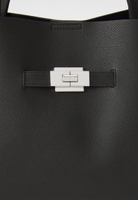 Svart skinn tote bag med teksturert finish, rektangulær spenne i sølv med "DKNY"-logo, og solide sorte håndtak.