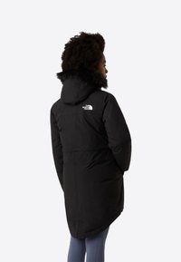 Μαύρο παρκά μπουφάν από μονωτικό ύφασμα, με κουκούλα επενδυμένη με τεχνητή γούνα και λευκό λογότυπο στο άνω πίσω μέρος. Μακριά γραμμή με ευθεία κοπή.