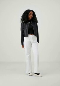 Donna con capelli ricci che indossa una giacca corta nera, un top nero, pantaloni bianchi a vita alta e sneaker bianco e nere, in piedi contro uno sfondo semplice.