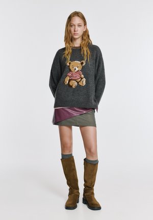 Grijze gebreide trui met een berenmotief, gedragen over een rok met pruimkleurige accenten. Bruine kniehoge laarzen met gespen, grijze decoratieve sokken.