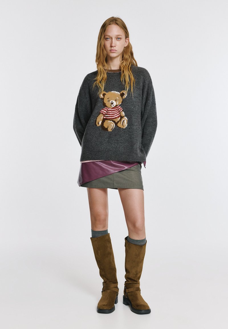 Szary dziergany sweter z motywem niedźwiedzia, noszony na spódnicy z akcentami w kolorze śliwkowym. Brązowe kozaki za kolano z klamrami, szare ozdobne skarpety.