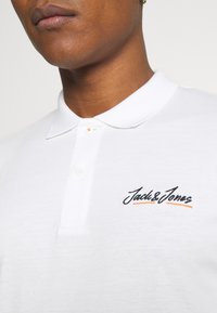 White polo shirt with a classic collar, featuring an embroidered "Jack&Jones" logo in black and orange on the chest. Button placket detail visible.