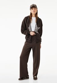 Sudadera con cremallera marrón con un diseño de estrella de patrón de leopardo, combinada con pantalones deportivos marrones holgados. El modelo lleva una camiseta blanca y una gorra marrón.