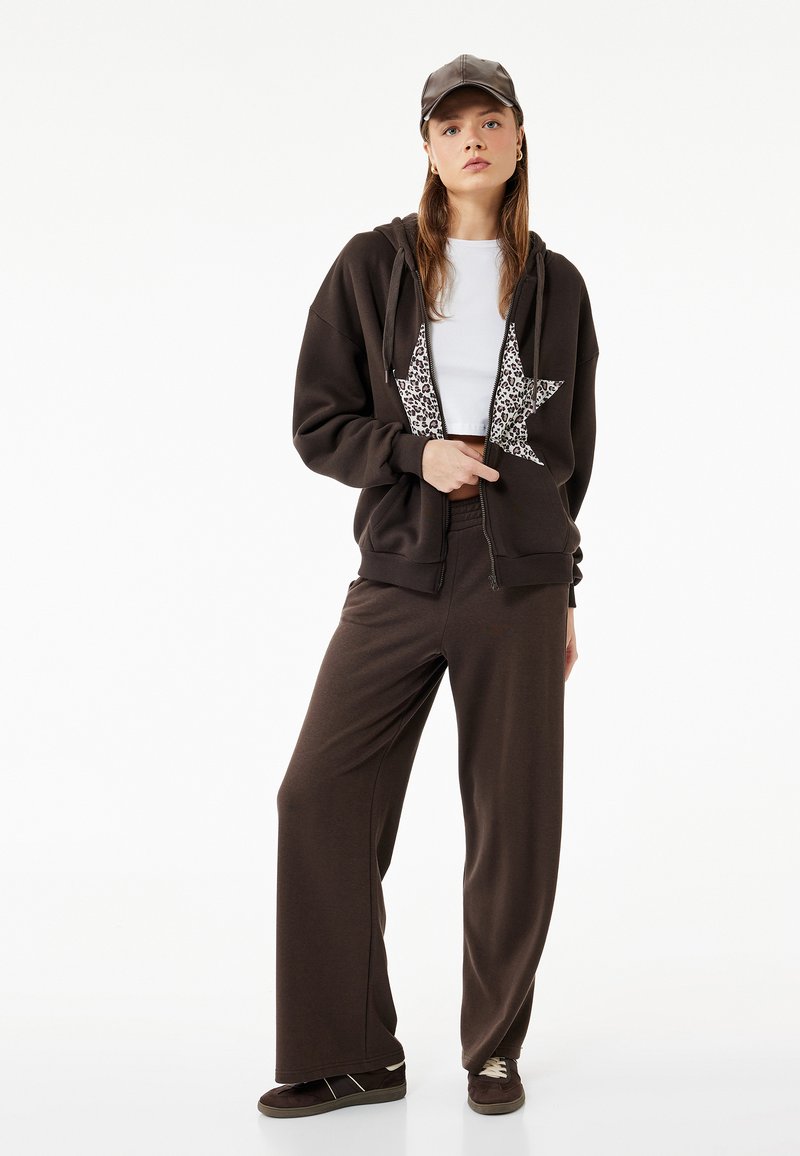 Sudadera con cremallera marrón con un diseño de estrella de patrón de leopardo, combinada con pantalones deportivos marrones holgados. El modelo lleva una camiseta blanca y una gorra marrón.