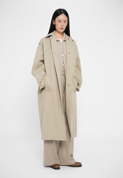 Μπεζ μακρύ παλτό φτιαγμένο από ύφασμα με γιακά και κούμπωμα, φορεμένο πάνω από ένα ανοιχτό καφέ τοπ και ταιριαστά παντελόνια με φαρδιά πόδια. Καφέ παπούτσια.