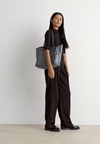 Sac fourre-tout en pied-de-poule noir fabriqué en matériau synthétique, avec deux poignées et une forme structurée. Le modèle porte une tenue entièrement noire.