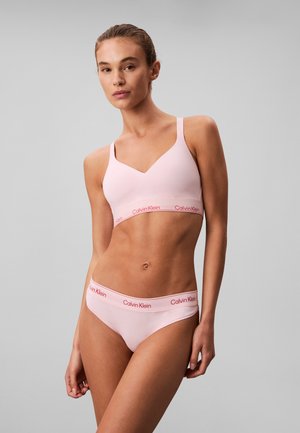 Femme portant un ensemble de sous-vêtements Calvin Klein rose clair sur un fond gris uni, debout avec une posture détendue et une expression neutre.