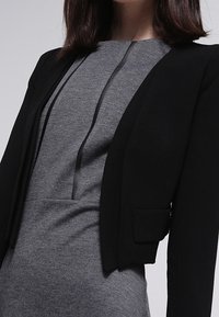 Svart kort blazer med en strukturerad passform, kombinerad med en grå klänning med rund halsringning och subtila detaljer fram. Slät tygtextur.