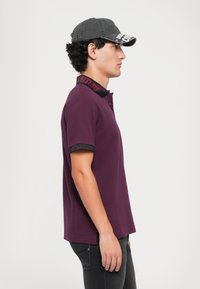 Jovem em uma camisa polo roxa escura com texto na gola, calças jeans pretas e um boné de beisebol preto, virado de lado em um fundo simples.