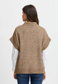 Brun strikket sweater med høj krave, korte ærmer og ribbet kant. Teksturen ser blød og komfortabel ud, lagdelt over en hvid skjorte.