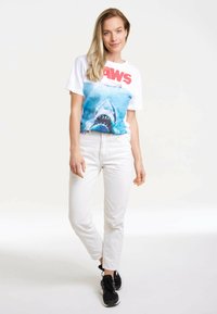 White short-sleeve t-shirt featuring a shark graphic and red text "JAWS"; paired with light-colored jeans and black sneakers.