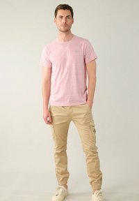 Hellrosa, kurzärmliges T-Shirt mit subtiler Textur. Kombiniert mit beigen Cargo-Hosen mit Seitentaschen und elastischen Bündchen. Weiße Sneaker.