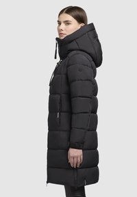 Abrigo acolchado negro hasta la rodilla con capucha, que presenta secciones acolchadas horizontales, cremalleras laterales y un parche con el logo en la manga.