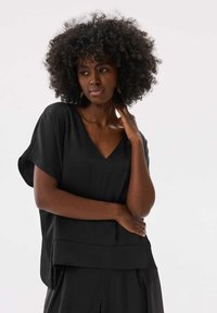 Zwarte blouse met een losse pasvorm, V-hals, korte mouwen en een gelaagde onderrand. Gladde stof met minimale details. Casual gedragen met gekruiste armen.