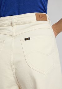 Los vaqueros de mezclilla color crema presentan un parche de cuero con la marca "Lee", costuras visibles y un bolsillo con un detalle de diseño curvado.