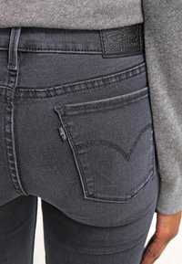 Jeans de mezclilla gris oscuro con una vista en primer plano del bolsillo trasero, que presenta una distintiva etiqueta de logo y un sutil detalle de costura.