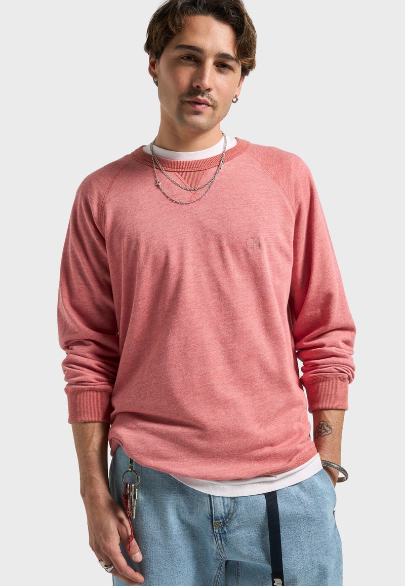 Sweatshirt à enfiler rose pâle avec manches raglan, poignets côtelés et un logo discret. Porté sur une chemise blanche, accompagné d'un pantalon en denim clair.