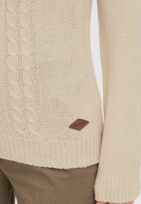 Beiger Strickpullover mit einem Kabelmuster, gerippten Bündchen und einem braunen Lederlogo-Patch an der Seite. Weiche Textur und lockere Passform.