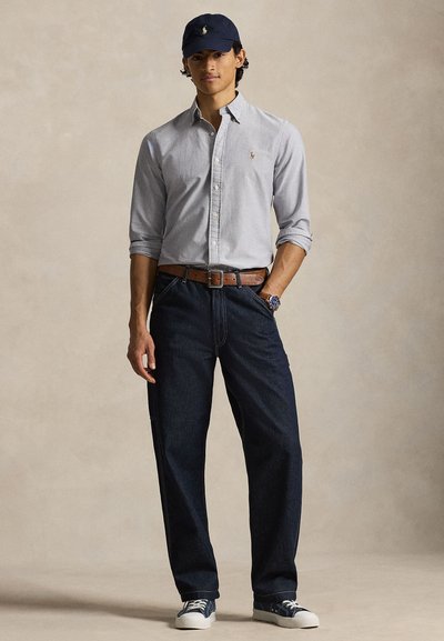 Young man wearing a light gray button-up shirt, dark blue jeans, navy cap, brown belt, blue sneakers, and a wristwatch, standing with one hand in pocket.