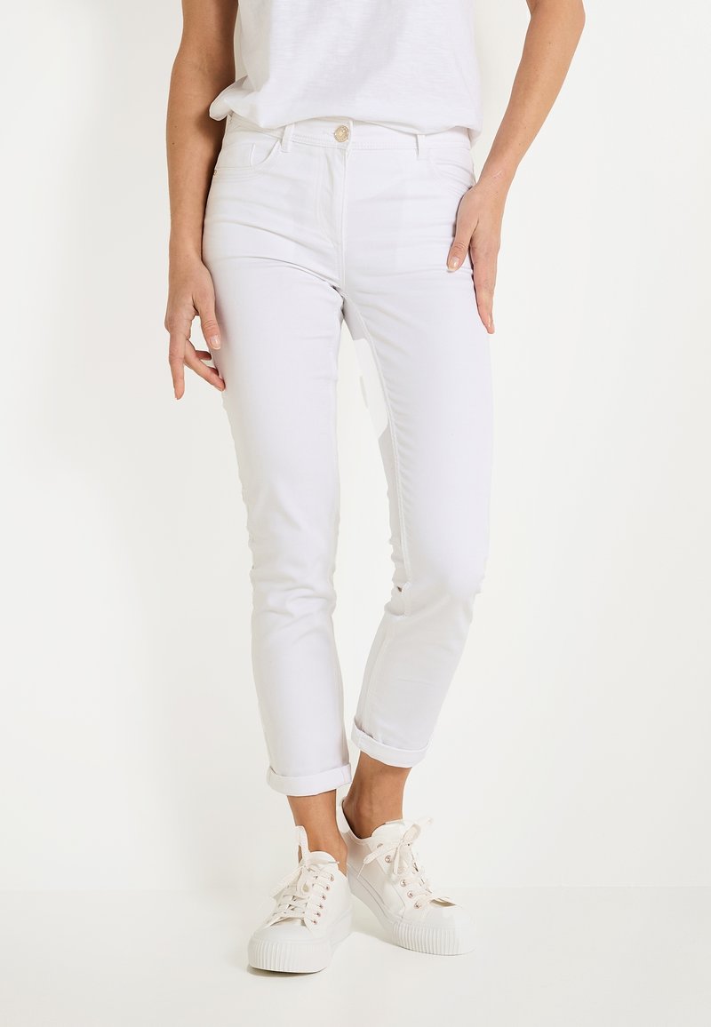 Vaqueros de mezclilla blancos de algodón, de longitud hasta el tobillo, ajustados y con puños enrollados, con cierre frontal de botón y cinco bolsillos. Llevados con zapatillas blancas.