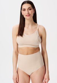 Kvinde med langt mørkt hår iført en beige amme-bh og matchende højtaljede trusser, står foran en simpel hvid baggrund.
