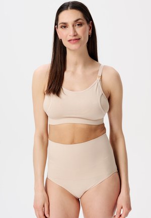Vrouw met lang donker haar, gekleed in een beige borstvoedingsbeha en bijpassende hoge onderbroek, staand tegen een effen witte achtergrond.