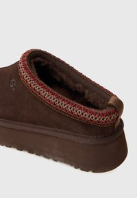 Mocassin en suede marron avec un intérieur doux. Il possède une bordure décorative en broderie rouge et marron le long de l'ouverture et une semelle en caoutchouc.