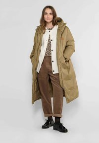 Khaki pufferjack met een capuchon, voorzien van een rits- en knoopsluiting, gedrapeerd over een crèmekleurige cardigan. Bruine corduroy broek en zwarte schoenen.