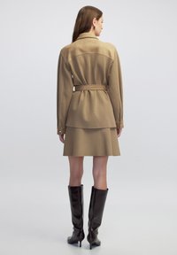 Vestido beige con cinturón, de mangas largas y falda en capas. Textura suave y diseño estructurado. Un par de botas marrón brillantes hasta la rodilla.