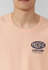 Licht perzik kleurige katoen t-shirt met een ronde halslijn, voorzien van een marineblauw ovaal logo met de tekst: "PACIFIC CUSTOMS BOARDS&KITS" gedrukt op de voorkant.