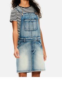 Vaaleansininen farkkuhaalari, jossa on etutaskut, säädettävät olkaimet ja istuva vyötärö. Yhdistetty mustavalkoraidallisen paidan kanssa.