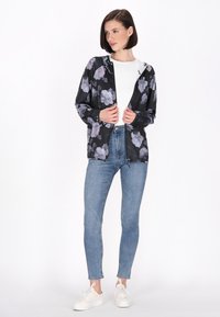 Giacca leggera nera con fiori viola, chiusura con zip, cappuccio, maniche lunghe, abbinata a jeans azzurri chiari e sneakers bianche.