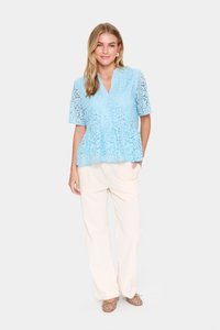 Šviesiai mėlynas nėriniuotas peplumas su trumpomis rankovėmis ir V formos iškirpte. Sujungtas su grietinėlės spalvos plačiomis kelnių ir neutraliomis atviromis bateliais.