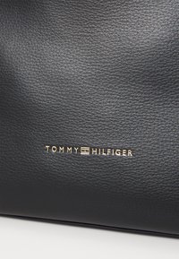 Musta nahkalaukku, jossa on teksturoitu pinta ja kultainen logo, jossa lukee "TOMMY HILFIGER", kaiverrettuna alaosaan.