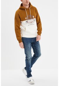 Sudadera marrón y blanca con logo hexagonal, con capucha ajustable y bolsillo frontal, combinada con vaqueros azules y zapatillas blancas.