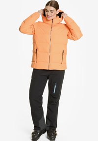 Chaqueta de plumón ligera color naranja con capucha, cierre de cremallera y dos bolsillos frontales con cremallera. Lleva unos pantalones negros con detalles en cremallera azul.
