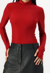 Rotes, langärmliges, hochgeschlossenes Oberteil aus glitzerndem, dehnbarem Material, kombiniert mit einem schwarzen Lederrock mit glatter Textur und taillierter Passform.